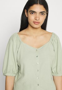 Blusa verde-clara com mangas bufantes, decote quadrado e duas linhas de botões, apresentando uma mistura de botões sólidos e transparentes. Textura suave.