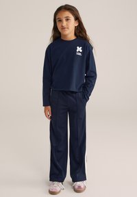 Jauna meitene ar gariem brūniem matiem, tērpusies tumši zila garām piedurknēm krekliņā un atbilstošās biksēs ar baltām sānu svītrām, valkā rozā sporta apavus.