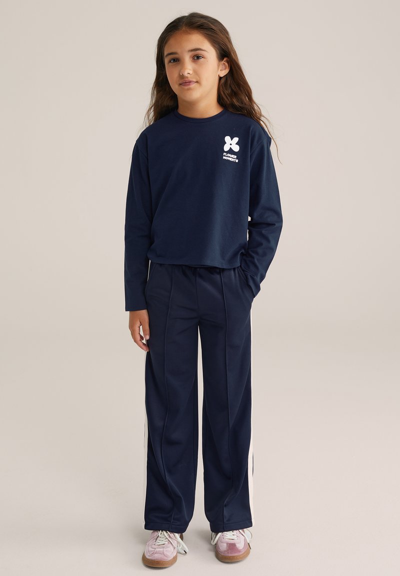 Jauna meitene ar gariem brūniem matiem, tērpusies tumši zila garām piedurknēm krekliņā un atbilstošās biksēs ar baltām sānu svītrām, valkā rozā sporta apavus.