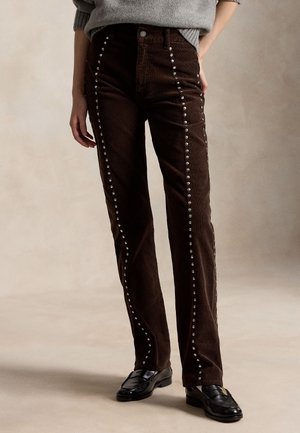 Persona che indossa pantaloni marroni in velluto a coste con borchie argentate verticali e mocassini neri, in piedi con una mano in tasca.