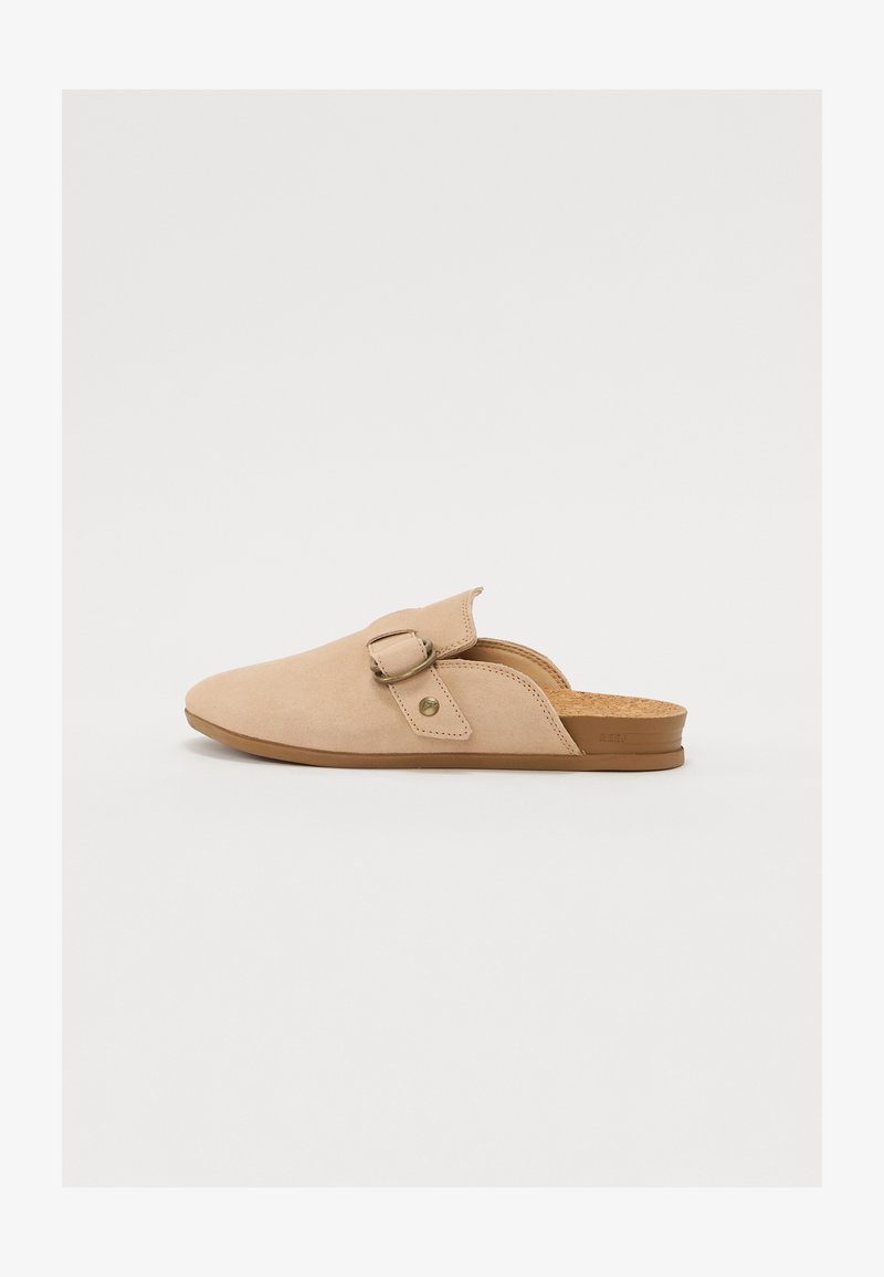 Beige suede slip-on shoe with a rounded toe, adjustable strap, and cork insole, featuring a minimal design and soft texture.