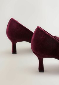 Pāris burgundijas krāsas samta zeķu papēžu ar kvadrātveida papēžiem uz gaiši pelēka fona.