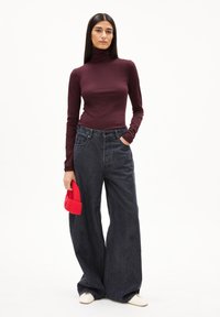 Burgunderfarbener Langarm-Turtleneck, kombiniert mit locker sitzenden dunklen Jeans, die mit einer Hand gehalten werden. Eine leuchtend rote Mütze wird mit der anderen Hand festgehalten.