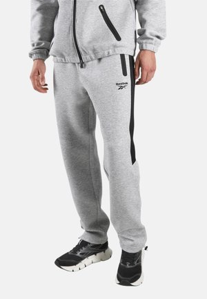 Man draagt grijze Reebok joggingbroek met zwarte zijstrepen en zwarte sport schoenen, staand tegen een witte achtergrond.