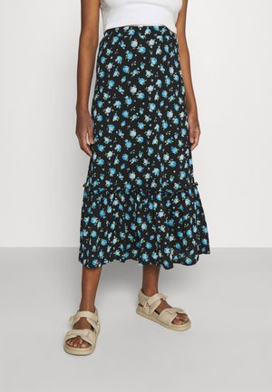 Femme portant une jupe midi noire à imprimé floral bleu, un haut blanc sans manches et des sandales ouvertes beiges avec des brides.