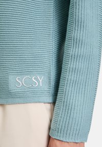 Pull en maille côtelée bleu clair avec un design court. Il présente une étiquette brodée blanche "SCSY" près de l'ourlet. Texture lisse.