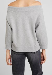 Sudadera gris sin hombros con un corte holgado, cuello y puños acanalados, y textura suave. Llevada con jeans oscuros, mostrando un estilo relajado.