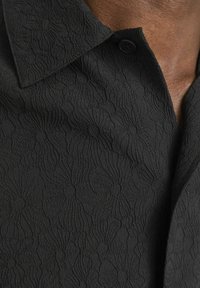 Zwarte gestructureerde shirt met bloemenpatroon, voorzien van een puntige kraag en een enkele zwarte knop bij de hals. Het materiaal lijkt zacht en gestructureerd.