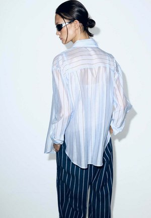 Mujer con el cabello oscuro recogido en un moño, llevando gafas de sol negras, una camisa blanca translúcida con rayas finas y pantalones azul marino con rayas finas, mirando de perfil.