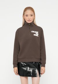Žena s rovnými blond vlasmi má na sebe hnedú mikinu na zips s bielym textom a lesklú čiernu mini sukňu s predným rozparkom.