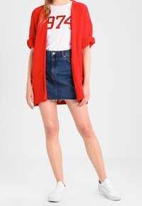 Czerwona, prążkowana kardigan z krótkimi rękawami na białym t-shircie z numerem "974", w połączeniu z niebieską spódnicą dżinsową i białymi tenisówkami.
