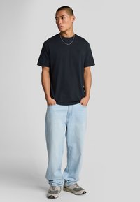 Camiseta de algodón negra con cuello redondo y mangas cortas, combinada con unos vaqueros de corte holgado en azul claro y zapatillas grises. Diseño simple.