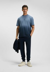 T-shirt a maniche corte in gradiente blu, con collo rotondo e logo. Abbinato a pantaloni jogger blu scuro e scarpe da ginnastica di colore chiaro.