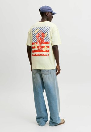 Homme portant un t-shirt jaune pâle avec un homard rouge, des tranches de citron et des rayures bleues et rouges dans le dos, un jean bleu clair et une casquette bleue.