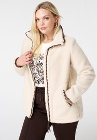 Blonde vrouw in een crèmekleurige fleecejas met bruine accenten, een wit shirt met patroon en donkerbruine broek, poseert met één hand in de zak en één hand bij de kraag.