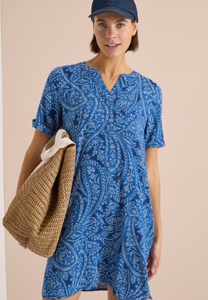 Kvinde iført en blå mønstret kjole og navy kasket, bærer en stor vævet taske over skulderen.