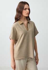 Polo a maniche corte color sabbia con righe orizzontali, dotato di colletto e scollatura a V. Tessuto morbido, vestibilità rilassata.