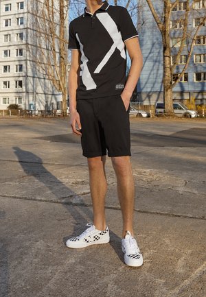 Mann steht draußen und trägt ein schwarz-weißes Poloshirt mit Grafik, schwarze Shorts und weiße Sneakers mit Schachbrettmuster.