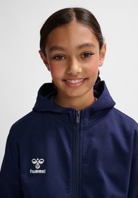 Dunkelblaue Zip-Hoodie aus weichem Stoff, mit Kapuze, slim Fit und einem weißen gestickten Logo auf der Brust.
