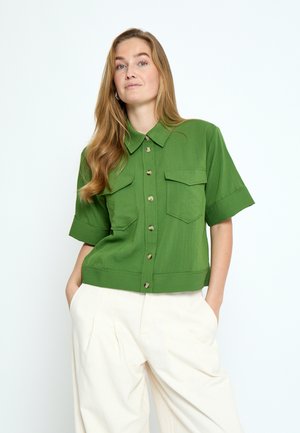 Frau mit langem Haar, die ein grünes kurzärmeliges Hemd mit Knöpfen und Taschen sowie lockere weiße Hosen trägt, Hände in den Taschen, nach vorne blickend.