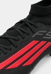 Μαύρο ποδοσφαιρικό παπούτσι με κόκκινες και γκρι ραβδώσεις, υφή στο πάνω μέρος και μαύρα κορδόνια. Ελαφρύς σχεδιασμός, ιδανικός για απόδοση στο γήπεδο.