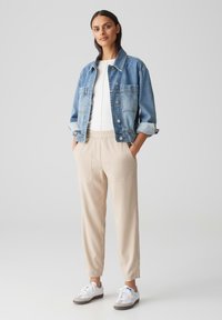 Denimjack met twee voorkruiken, beige elastische taillebroek en witte sneakers met grijze accenten. Model staat tegen een effen achtergrond.