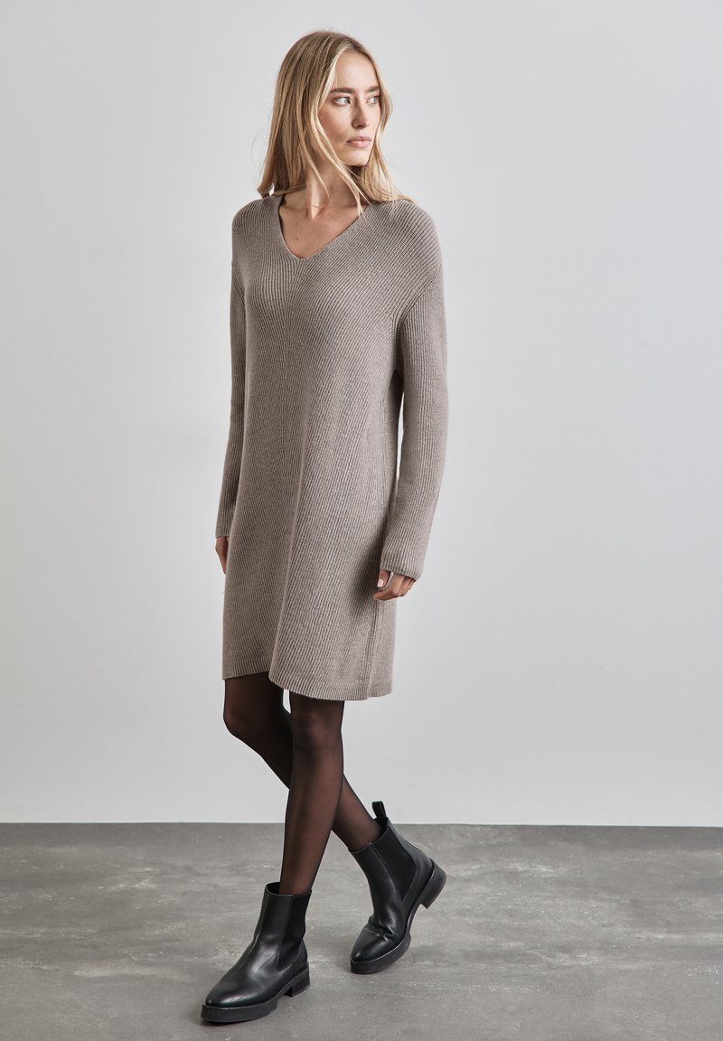 Strickkleid inTaupe mit V-Ausschnitt, langen Ärmeln und gerippter Textur, kombiniert mit schwarzen Knöchelstiefeln und transparenten Strumpfhosen. Minimalistisches Design.