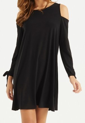 Robe noire mi-longue avec découpes aux épaules et manches longues nouées aux poignets, portée par une femme aux cheveux ondulés.