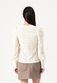 Kvinna med långt mörkt hår bär en vit texturerad långärmad blus med puffiga axlar och en beige mockajerkortkjol, stående mot en vit bakgrund.