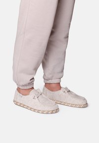 Lichtgrijze vilten pantoffels hebben een ronde neussluiting, een decoratieve strik en een gevlochten zool. Gekleed met aansluitende beige trainingsbroek.