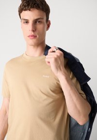 Beige kokvilnas T-krekls ar apaļu kakla izgriezumu un HUGO logo. Modelis tur tumšu džinsu jaku pār pleca, ar ziliem akcentiem.