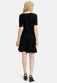 Schwarzes kurzärmeliges Kleid mit ausgestellt Rock. Aus weichem Stoff gefertigt, mit einem runden Ausschnitt im Rücken und knieumspielendem Saum. Trägt schwarze Absatzsandalen.