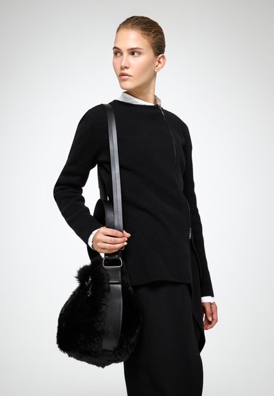 Maglione nero a maglia con zip frontale, indossato sopra una camicia bianca con colletto. Una borsa in pelliccia nera a trama con una tracolla in pelle è tenuta nella mano sinistra.