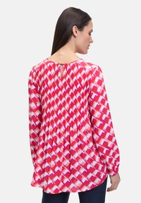 Blusa a maniche lunghe con retro plissettato, motivi geometrici rosa e rossi, e chiusura a chiave nel collo.
