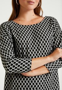 Schwarz-weiß gemusterte Bluse mit rundem Halsausschnitt, dreiviertel Ärmeln und einem geometrischen Design mit Dreiecken und Zickzackmotiven.