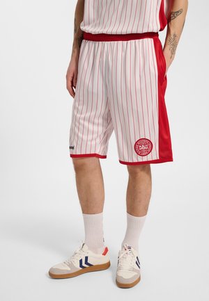 Man draagt witte sportshorts met rode verticale strepen en rode zijpanelen, met een ronde rode logo en witte sneakers met blauwe accenten.