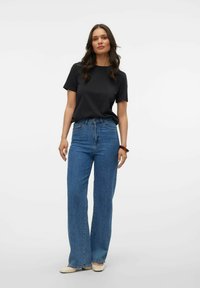 Schwarzes T-Shirt mit kurzen Ärmeln kombiniert mit hochgeschnittenen, weit geschnittenen blauen Jeans. Das Model trägt beigen Schuhe und ein braunes Armreif.