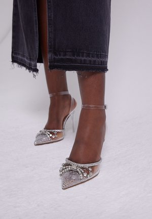 Sølv høje hæle med spidse tæer, der har klare ankelremme, perleudsmykninger og en glitrende tekstureret overflade.