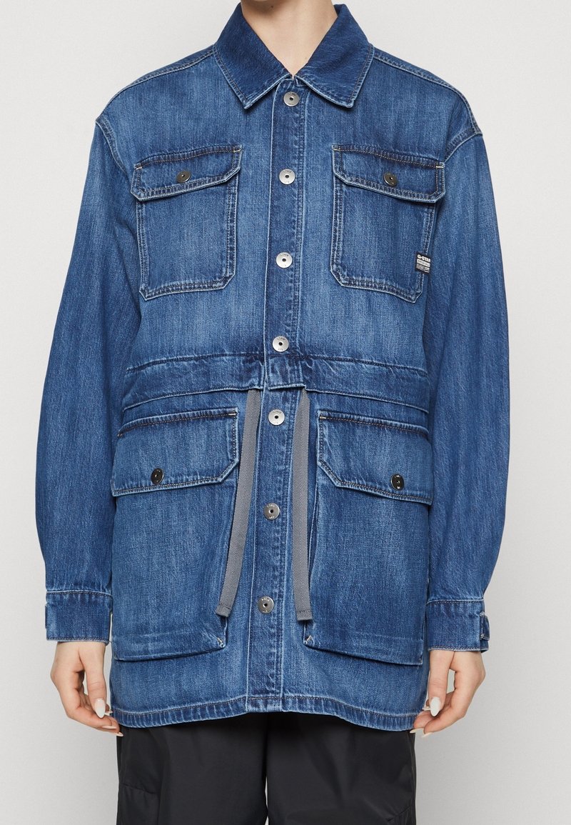 Veste en jean bleue à manches longues avec quatre poches à rabat boutonnées et cordon gris à la taille portée par une personne.