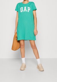 Teal T-Shirt-Kleid mit weißem "GAP" Logo, kurzen Ärmeln, lockerer Passform. Kombiniert mit einer beigen Handtasche und beige-farbenen, robusten Slippern. Weiße Socken sichtbar.