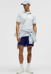 Jasnoniebieska koszulka polo, niebieskie szorty, białe sneakersy i niebieska czapka. Sweter związany w pasie. Gładkie tekstury, swobodny styl.