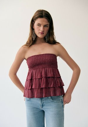 Jeune femme portant un top bordeaux sans bretelles et à volants avec des rayures subtiles, un jean bleu clair et de grandes boucles d'oreilles géométriques, sur un fond uni.