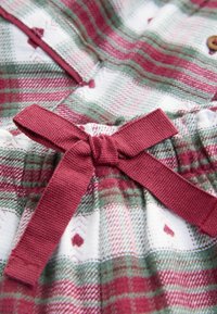 Rot-grüner Karostoff mit einer rosa Schleife an der Taille. Das Material wirkt weich mit einer strukturierten Oberfläche und kleinen geometrischen Mustern.