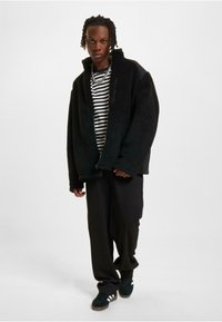 Schwarze Fleecejacke mit hohem Kragen und Reißverschluss-Brusttasche, getragen über einem schwarz-weiß gestreiften Hemd. Schwarze, locker sitzende Hose und Schuhe.