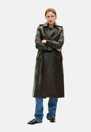 Kvinde iført lang, mørkebrun læder trenchcoat, blå jeans og sorte spidse flade sko, stående med armene krydset mod hvid baggrund.