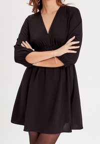 Robe noire en tissu texturé, col en V et manches trois-quarts bouffantes. Taille cintrée menant à une jupe évasée.
