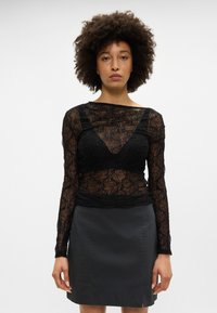 Čierna čipkovaná blúzka s dlhými rukávmi a výstrihom do V, zdobená perlovými detailmi a viacerými textúrami. Doplní ju priliehavá čierna sukňa.