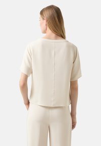 Kurzärmlige Bluse aus leicht beigem Stoff, mit einem runden Halsausschnitt, geradem Schnitt und einer Nahtdetail, die entlang der Mitte des Rückens verläuft.
