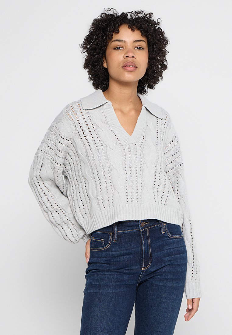 Pullover grigio chiaro a maniche corte con scollatura a V e colletto, caratterizzato da motivi a treccia e dettagli di cucitura aperta. Indossato con jeans scuri.
