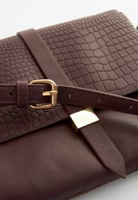 Sac à main en cuir marron avec texture gaufrée crocodile, boucle en matériel doré et une bandoulière plate, mettant en valeur un design raffiné et une surface lisse.